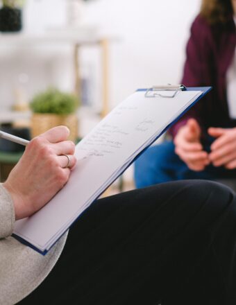 Close-up of a therapist writing notes on a clipboard while conversing with a patient.